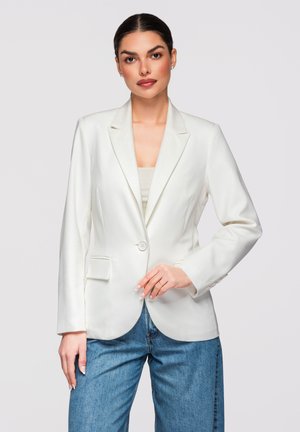 Žena v bílém jednokřídlém saku přes béžový top a modré džíny, pózuje před jednoduchým světle šedým pozadím.