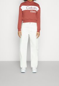 Sudadera roja con panel blanco que presenta el logo de "Calvin Klein", combinado con jeans blancos de corte holgado y zapatillas blancas. Textura de tela suave.