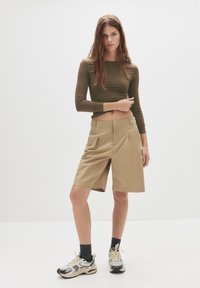 Brun langeærmet top parret med beige skræddersyede shorts. Sneakers har et mesh- og metaldesign. Modellen står foran en ensfarvet baggrund.