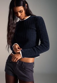 Temno modra pulover iz rebrastega pletenja z okroglim izrezom, obrobljen z belo kontrastno obrobo, kombiniran s temno sivimi jeans kratkimi hlačami. Gladek tekstura, oprijet kroj.