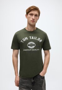 Σκούρο πράσινο βαμβακερό t-shirt με το κείμενο "TOM TAILOR" τυπωμένο και έναν κυκλικό θυρεό. Κοντά μανίκια και λαιμόκοψη τύπου crew neck. Άνετη εφαρμογή.