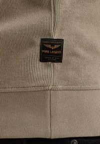 Tissu tricoté côtelé beige avec une étiquette rectangulaire noire portant l’inscription "PME Legend American Classic Tested at High Altitude" en texte doré.