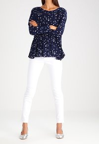 Maglietta a maniche lunghe blu navy con motivo a stelle bianche, design peplum; abbinata a pantaloni bianchi attillati e scarpe con tacco alto argentate.