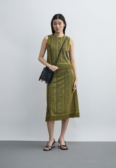 Γυναίκα με αμάνικο φόρεμα με ελαιώδες πράσινο σχέδιο και κρόσσια στο τελείωμα, μαύρη τσάντα χιαστί και μαύρα πέδιλα με λουράκια, που στέκεται μπροστά σε γκρίζο φόντο.