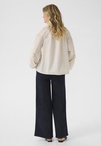 Blouse en lin beige avec des manches volumineuses, broderie détaillée sur les poignets, associée à un pantalon large bleu marine et des sandales noires.
