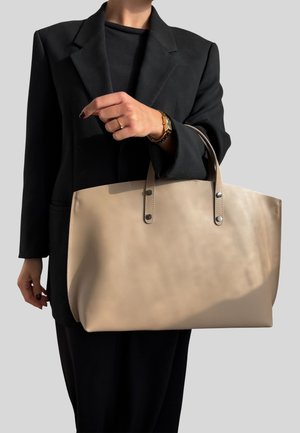 Borsa a tracolla in pelle beige con manici superiori, design minimalista, texture liscia, posizionata su uno sfondo grigio; modello che indossa un blazer nero.