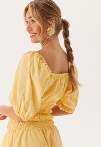 Gul bluse med puffærmer, firkantet halsudskæring og elastisk talje. Glat stof med en samlet tekstur. Modellen har håret i en hestehale.