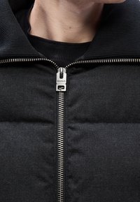 Gros plan sur une personne portant une veste matelassée noire avec une fermeture éclair argentée de marque Hugo et un col côtelé haut.
