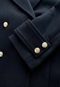 Tengerészeti gyapjú kabát részlete, széles mandzsettával, arany tónusú díszgombokkal és finom halszálkás textúrával.