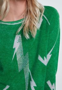 Pull vert avec des motifs d'éclairs blancs et des accents de strass argentés. Fabriqué en matériau doux et texturé avec un col décontracté.