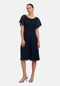 Robe bleu marine avec des manches flottantes transparentes, coupe mi-longue, taille cintrée et corsage plissé. Portée avec des talons argentés à straps.