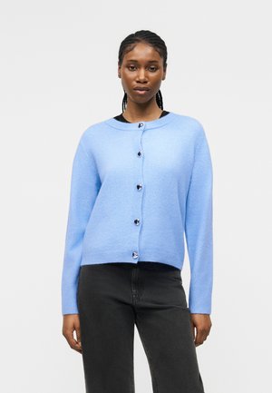 Frau mit geflochtenem Haar, die eine hellblaue Strickjacke mit herzförmigen Knöpfen und schwarze Hose trägt, steht vor einem weißen Hintergrund.