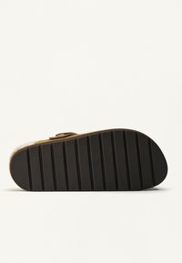 Hnedé sandále s hrubou čiernou podrážkou so zúbkami, viditeľným prackovým popruhom a krémovou plyšovou podšívkou na bielom pozadí.