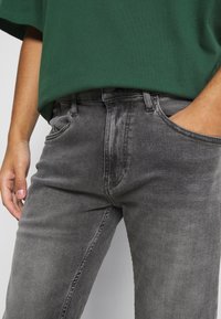 Vaqueros de mezclilla grises con corte ajustado, que cuentan con un cierre de botón, bolsillos frontales y una sutil textura desgastada. Estilizados con una blusa verde.