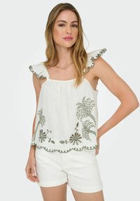 Top blanco sin mangas con tirantes con volantes, con bordado floral verde y dobladillo ondulado. Se lleva con pantalones cortos blancos.