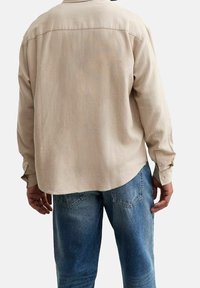 Beige pitkähihaispaita, jossa on suorakaiteen muotoinen leikkaus, napilliset kalvosimet ja takana olkapäämyssy, yhdistetty vaaleansinisiin farkkuihin, joissa on takataskut.