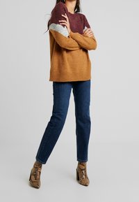 Stickad tröja med färgblockdesign: vinröd, grå och senapsgul. Mjuk textur, lös passform och ribbad nederkant. Matchad med mörkblå jeans och klackar med ormtryck.