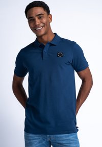 Námořnická modrá polo košile z bavlny, s malým černým emblémem na hrudi, krátkými rukávy a klasickým límcem.