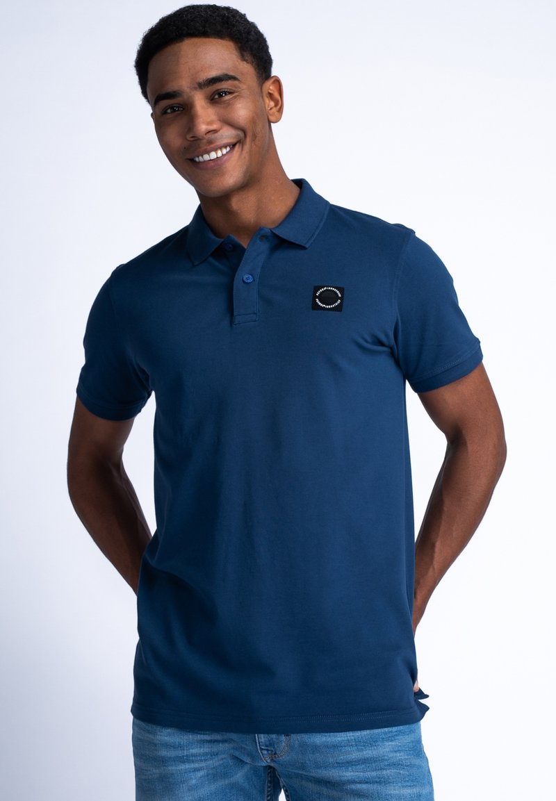 Námořnická modrá polo košile z bavlny, s malým černým emblémem na hrudi, krátkými rukávy a klasickým límcem.
