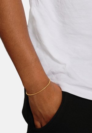 Bracelet en chaîne en or au poignet, présentant une surface lisse et brillante avec des maillons entrelacés. Porté avec une chemise blanche et un pantalon noir.
