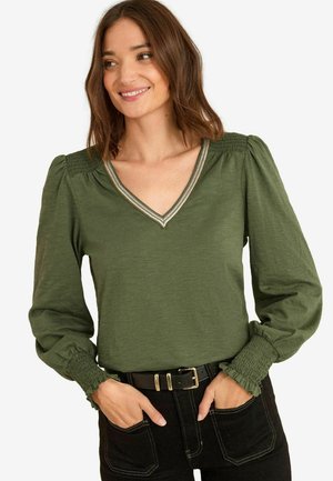 Woman wearing an olive green long-sleeve blouse with smocked shoulders and cuffs, paired with black pants and a black belt.