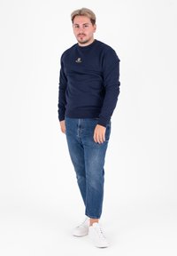 Granatowy sweter crewneck z złotym logo, w połączeniu z jasnoniebieskimi dżinsami i białymi trampkami. Prosty design, miękka faktura i luźny krój.