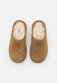 Pantofole slip-on in suede marrone con un morbido rivestimento interno in ecopelliccia crema. Il design presenta una punta arrotondata e bordi rinforzati.