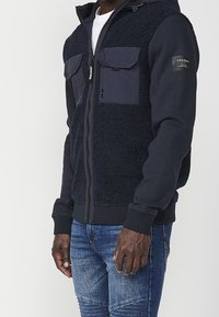 Marinblå zip-up hoodie med fleece och tygpaneler, med bröstfickor, ribbade ärmslut och justerbar huva. Texturerat material.