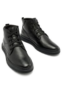 Paire de bottines en cuir noir à surface texturée, lacets noirs, et semelles épaisses en caoutchouc, présentées sur un fond blanc.