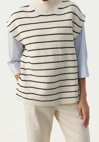 Ärmellose cremefarbene Strickweste mit schwarzen horizontalen Streifen, getragen über einem hellblauen Langarmshirt und cremefarbigen Pants, Hände entspannt an den Seiten.