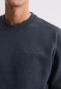 Ciemnoniebieska bluza z okrągłym dekoltem i subtelnym haftem logo w jaśniejszym kolorze. Wykonana z miękkiego, teksturowanego materiału, z rękawami raglanowymi.