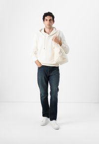 Krémová mikina sšnúrkami, tmavomodré džínsy a biele tenisky. Modelka drží svetlú tašku. Jednoduchý, nenáročný outfit.