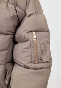 Manica di giacca imbottita in marrone tenue con una cerniera brillantinata color oro rosa. Presenta una texture trapuntata e una tasca aggiuntiva con dettaglio a soffietto.