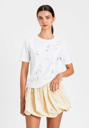 Giovane donna con capelli scuri raccolti in una coda di cavallo indossa una maglia bianca a maniche corte e una gonna beige chiaro con decorazioni di paillettes.