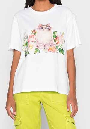 Frau trägt ein weißes T-Shirt mit einem sitzenden, flauschigen Kater, umgeben von pinken und gelben Blumen, die vorne aufgedruckt sind.