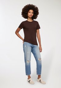 Maglietta corta marrone, jeans light blue strappati con buchi, e scarpe da ginnastica beige con dettagli neri, in piedi davanti a uno sfondo neutro.