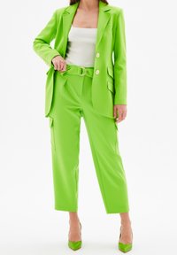 Neon groene pak met een getailleerde blazer en hoge taille broeken. De blazer heeft een knoopsluiting en zakken; de broeken hebben zijcargo zakken.