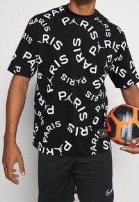 Mężczyzna w czarnych szortach i czarnej koszulce z rozsianym białym napisem "PARIS", trzymający piłkę nożną w czarno-pomarańczowych kolorach.