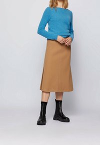 Femme portant un pull bleu, une jupe midi couleur camel avec une fente sur le côté, et des bottines noires se tenant devant un fond clair.
