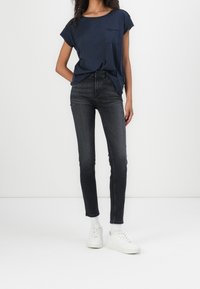 Námořnická modrá tričko s krátkým rukávem a kapsou, kombinované s tmavými úzkými džíny a bílými teniskami. Jednoduchý design, hladká textura a přiléhavý střih.