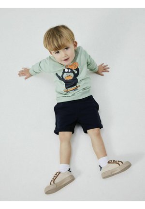Camisa de manga larga verde claro con un gráfico de un oso de dibujos animados y una pelota de baloncesto, combinada con pantalones cortos azul marino y zapatillas beige con acentos marrones.