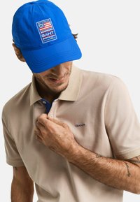Beige polo-skjorte med korte ermer og en blå knappestolpe, kombinert med en blå caps med logo og stripete detaljer.