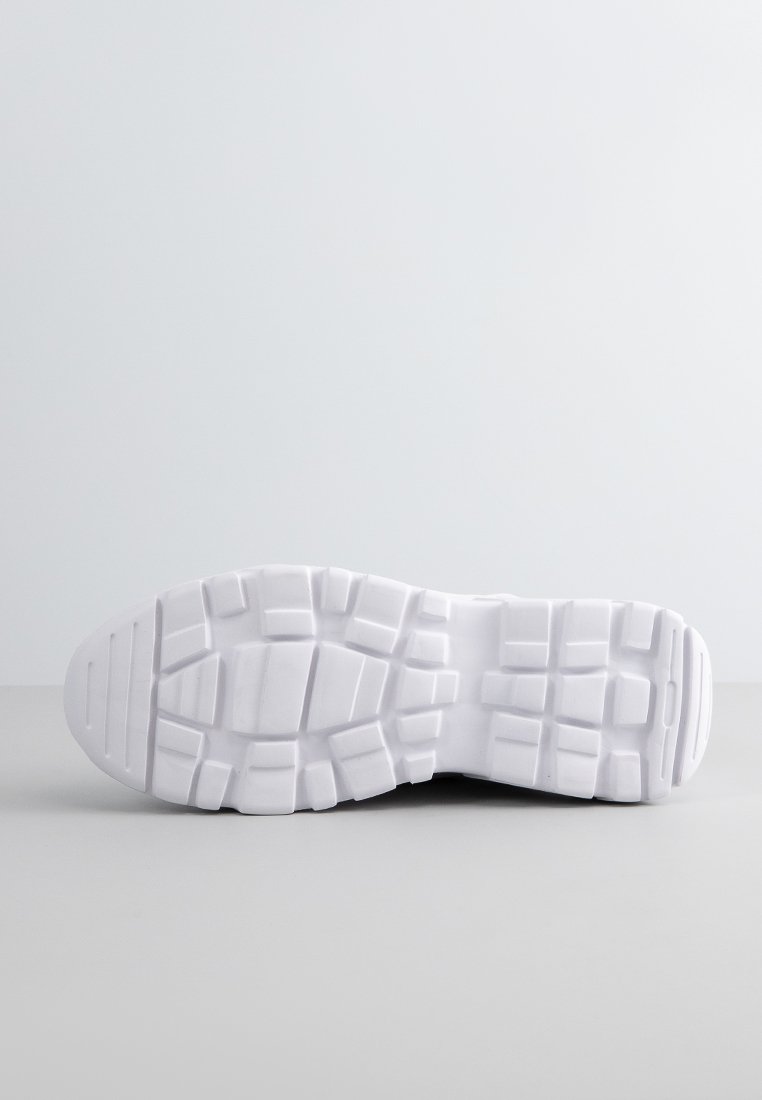 Suela de una zapatilla blanca de goma con surcos profundos y patrones geométricos, descansando sobre una superficie gris claro contra un fondo gris liso.