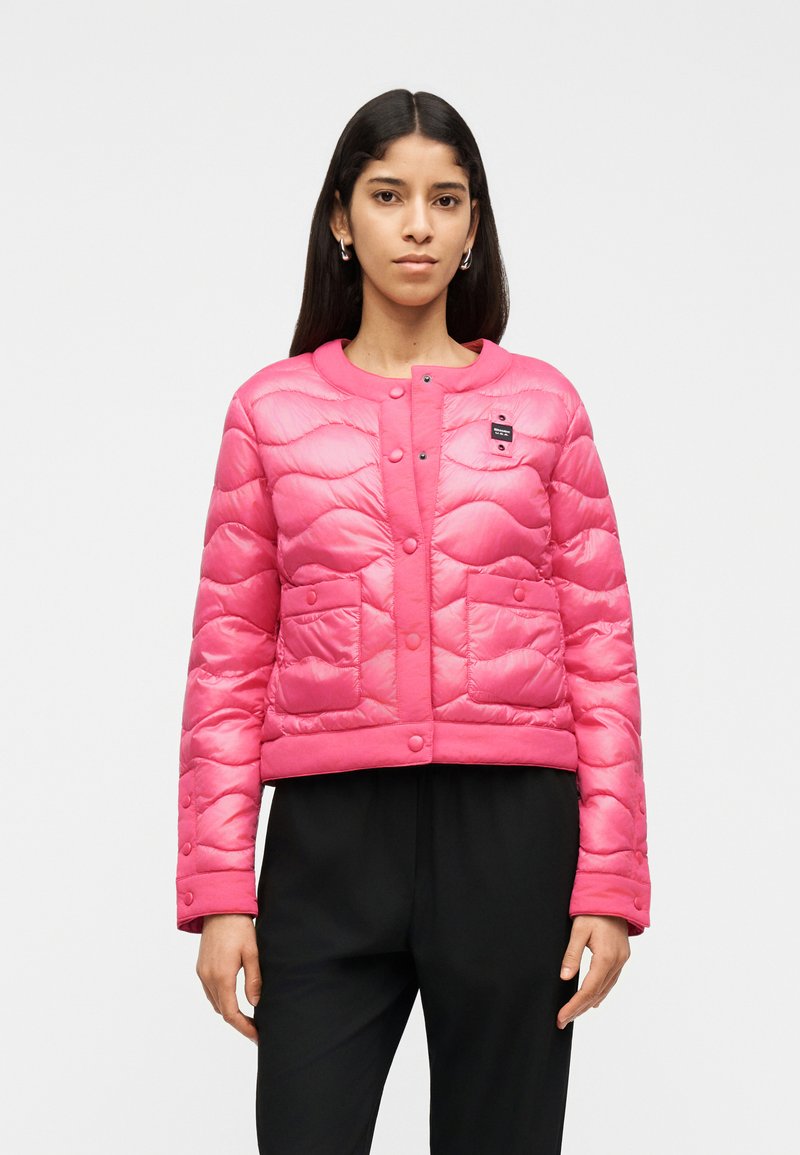 Γυναίκα που φοράει ένα έντονο ροζ καπιτονέ μπουφάν με κουμπιά τύπου snap, δύο εμπρόσθιες τσέπες και μαύρο παντελόνι, στέκεται μπροστά σε ένα απλό λευκό φόντο.