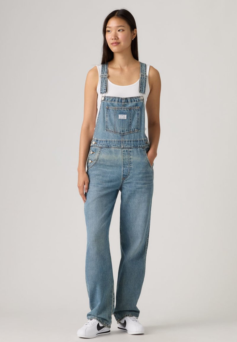 Φωτεινές μπλε τζιν σαλοπέτες με φαρδιά ποδόγυρα, ρυθμιζόμενες τιράντες, μπροστινή τσέπη και πλευρικά κουμπιά, συνδυασμένες με λευκή αμάνικη μπλούζα και λευκά αθλητικά παπούτσια.