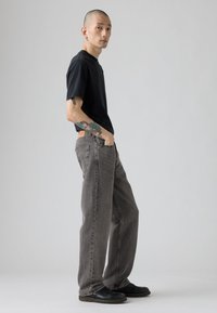 Crna majica kratkih rukava uparena s širokim sivim trapericama. Traperice imaju izblijedjeli izgled i zakrpu iznad stražnjeg džepa. Crne cipele.