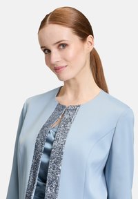 Jasnoniebieska kurtka z teksturowanym srebrnym haftem wzdłuż otwarcia z przodu. Gładka tkanina z klasycznym dekoltem i długimi rękawami.