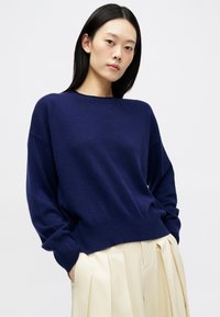 Jeune femme aux cheveux noirs raides portant un pull ample bleu marine et un pantalon large plissé crème, les mains dans les poches, fond neutre.