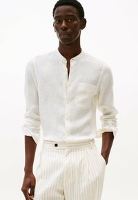 Camisa de lino blanca con cuello mandarín, botones en el frente, mangas largas arremangadas en los puños y un único bolsillo en el pecho.