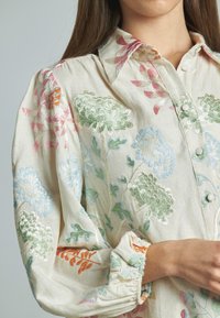 Camicia button-up ricamata di un leggero beige, con motivi floreali multicolori in verde, rosa e blu. Polsini arricciati e tessuto strutturato.